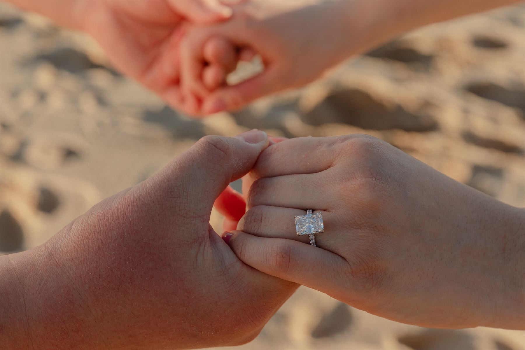 A couple holding hands on the beach, an engagement ring catching the light